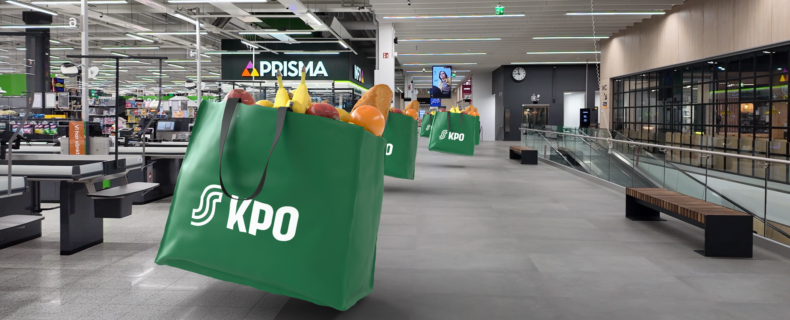 Inside a supermarket, large green reusable bags filled with groceries hang along a wide walkway with a Prisma store sign in the background.