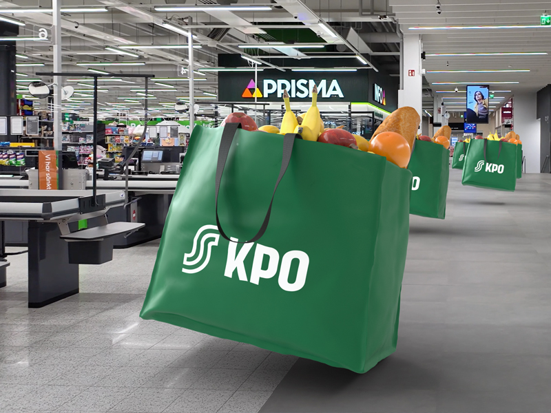 Green reusable bag with the KPO logo filled with groceries at a supermarket checkout area, blurred background of tills and Prisma sign.