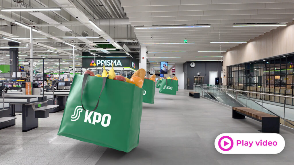 Inside a modern supermarket, large green reusable bags filled with groceries float in the foreground with a Prisma store sign in the background.