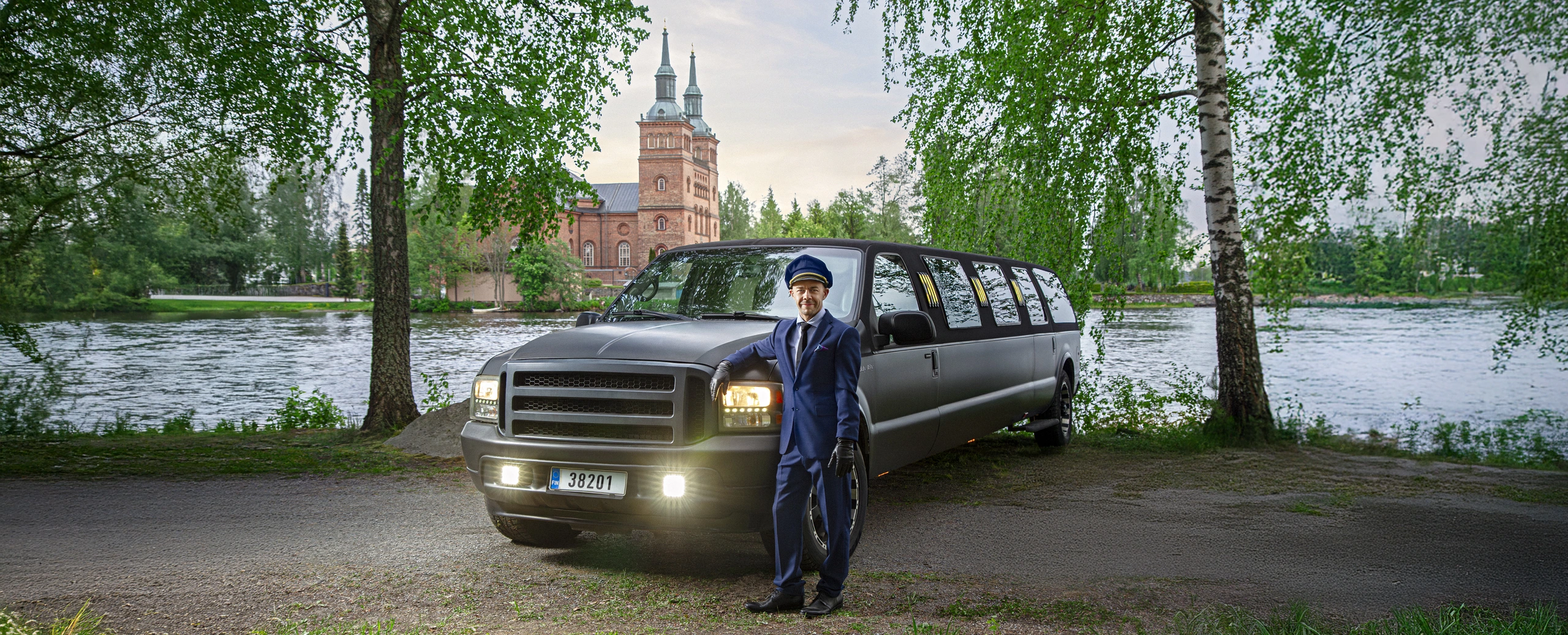Uniformed chauffeur leaning on a black stretch limousine beside a river, with a brick church and trees in the background.