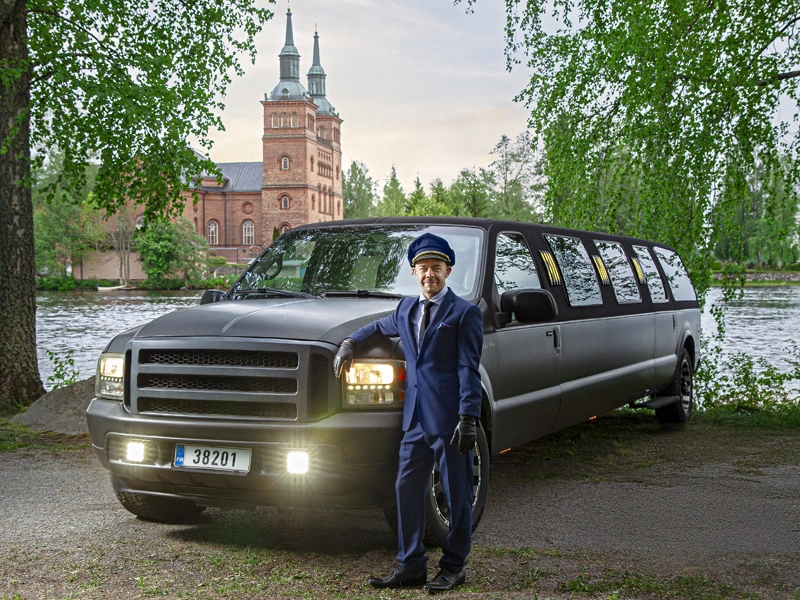 Chauffeur in a navy uniform stands beside a dark grey passenger van by a lakeside with a brick church in the background.
