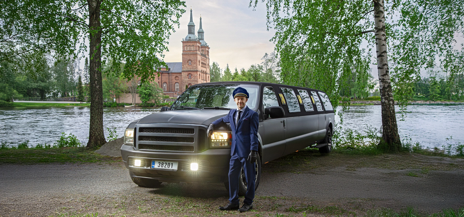 Chauffeur in a navy uniform stands beside a black stretch limo parked by a lake with a brick church in the background.