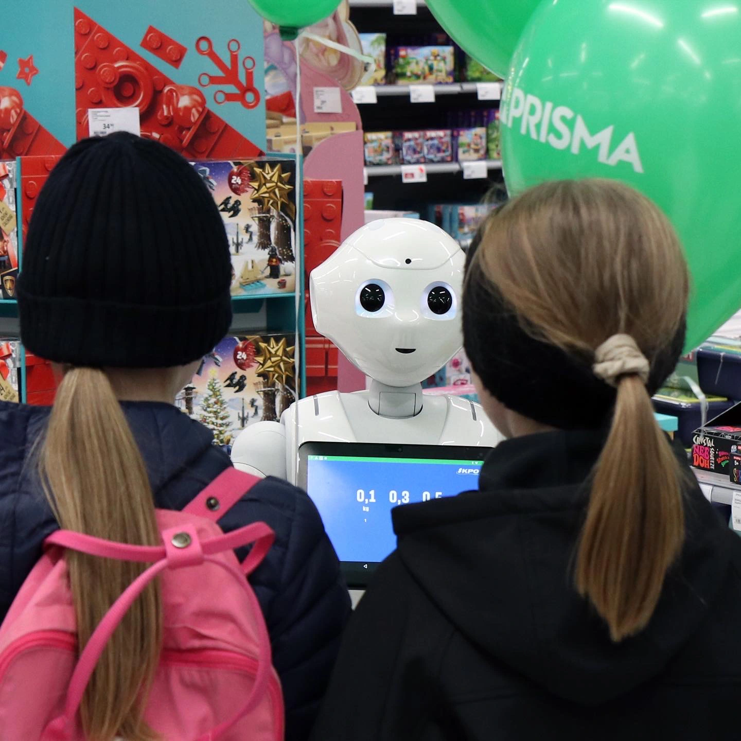 Two children with backpacks face a white friendly robot at a store kiosk in a retail setting.