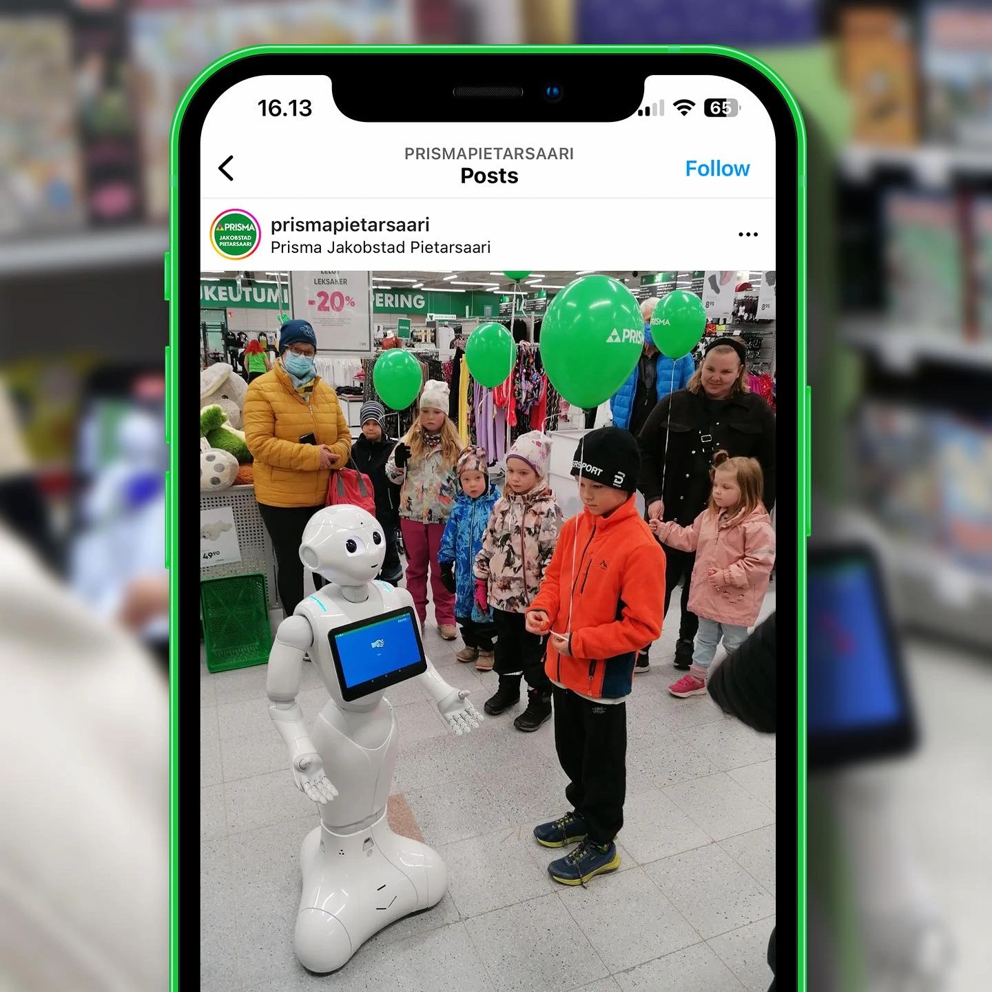 White humanoid robot entertaining a group of children in a store, with green balloons and shoppers in the background.