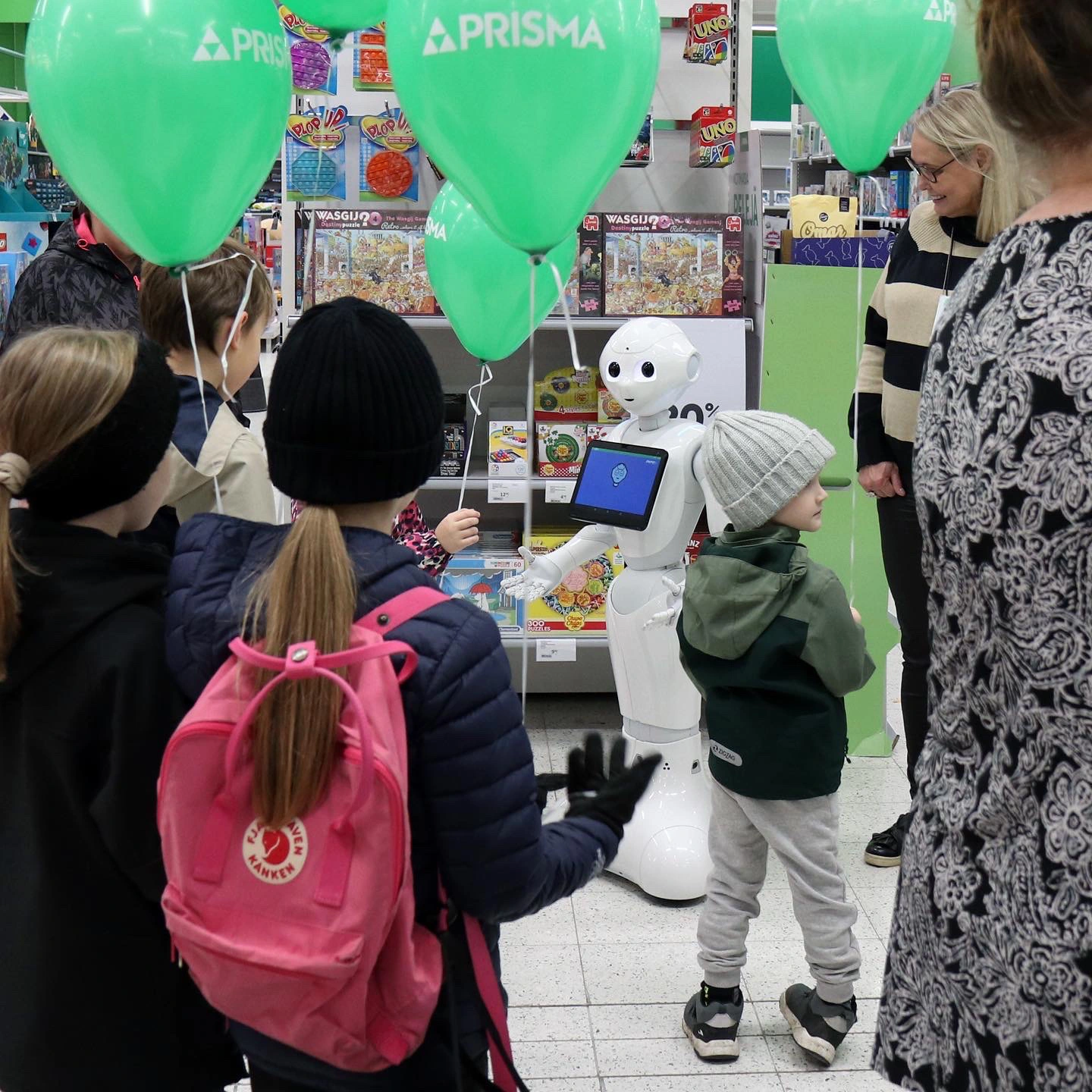 White humanoid robot with a tablet on its chest interacts with children in a toy store, with green balloons overhead as kids watch.