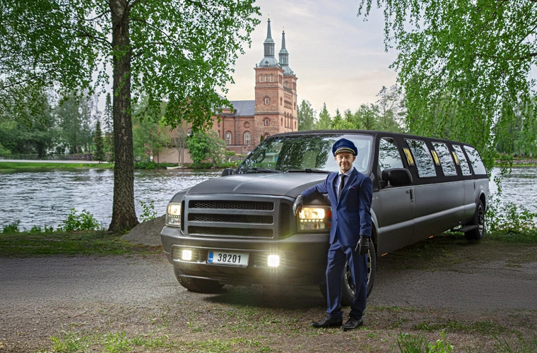 Chauffeur in a navy uniform and cap standing beside a long black limousine parked by a lake with a brick church in the background.